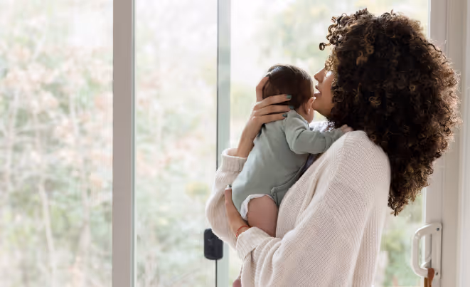 Mother holding and kissing infant by bright window with natural light streaming in, showing tender moment of affection.