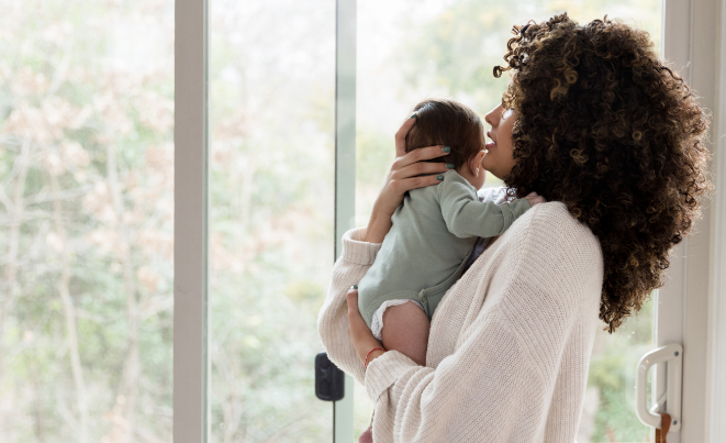 Mother holding and kissing infant by bright window with natural light streaming in, showing tender moment of affection.