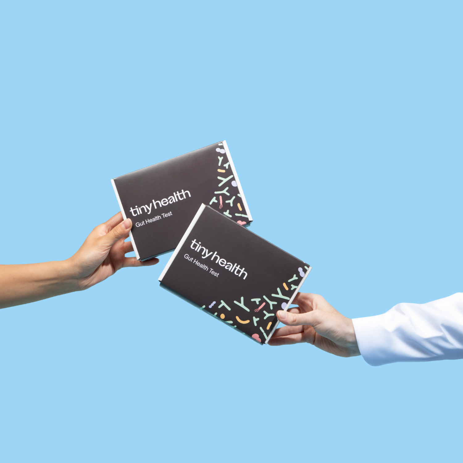 Two hands hold Tiny Health gut health test kits side by side, one arm in a lab coat, on a light blue background.