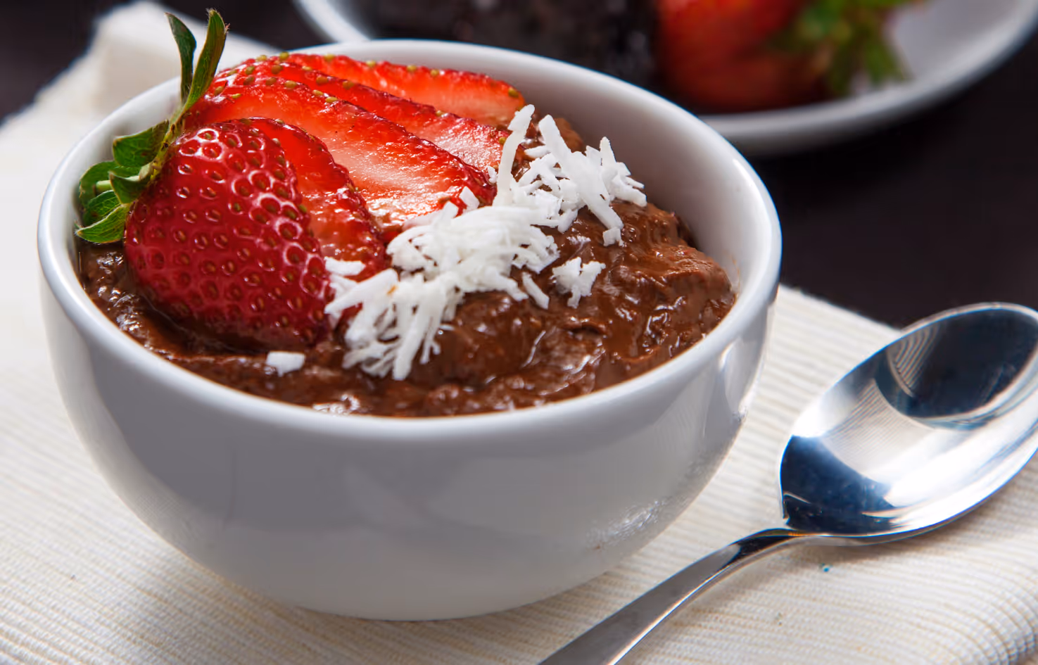 Chocolate avocado mousse in a white bowl topped with sliced strawberry and shredded coconut, with berries in background