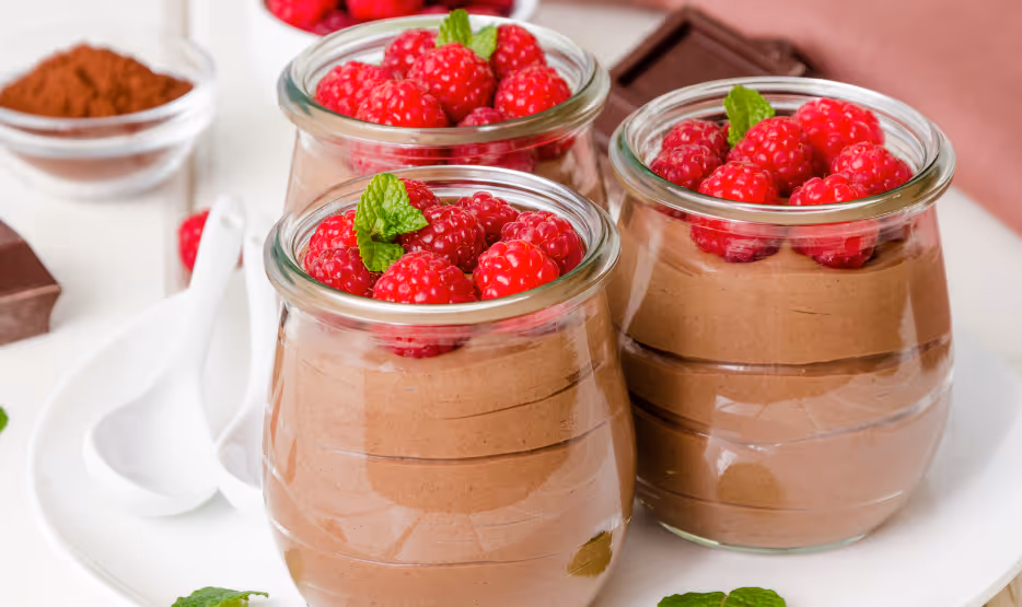 Three chocolate chia mousse servings in glass jars topped with fresh raspberries and mint leaves on white