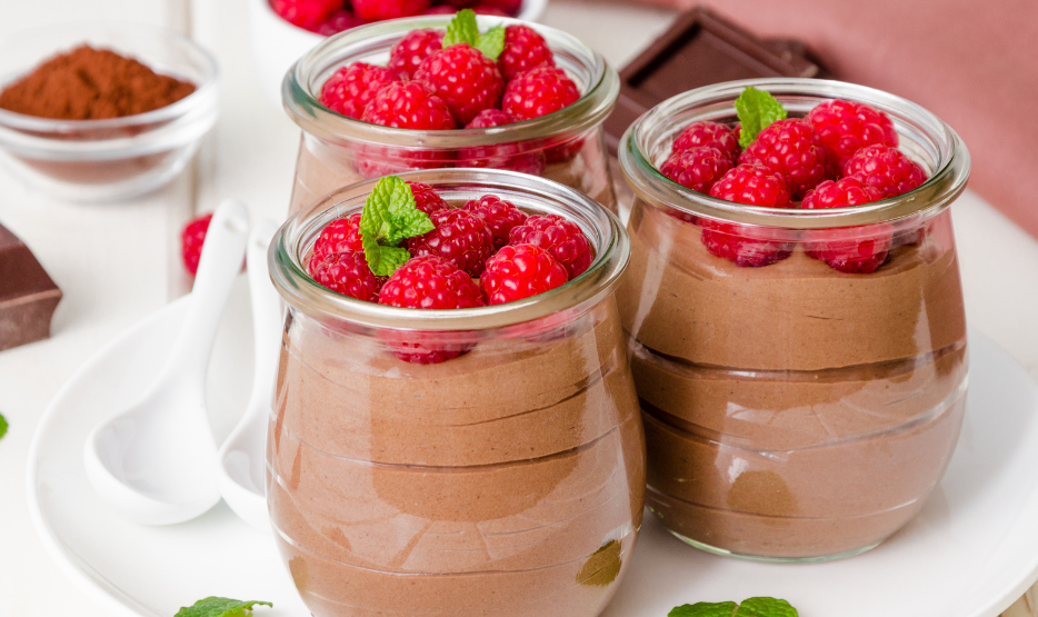 Three chocolate chia mousse servings in glass jars topped with fresh raspberries and mint leaves on white
