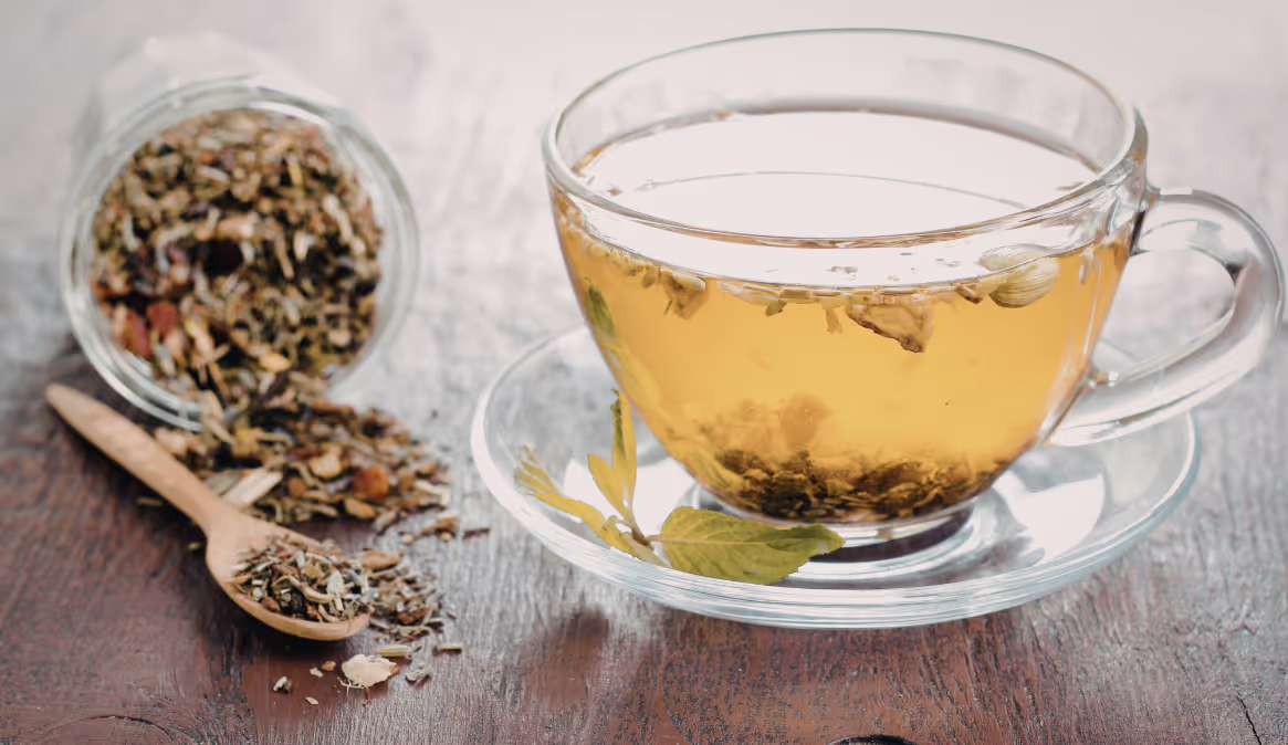 Glass cup of herbal tea on saucer with loose leaf herbs and a wooden spoon on a rustic wooden surface.