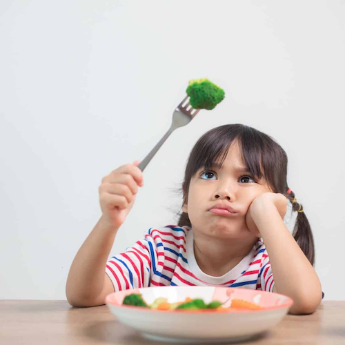 Young girl holds up a fork with broccoli, looking unimpressed, with a plate of vegetables in front of her.