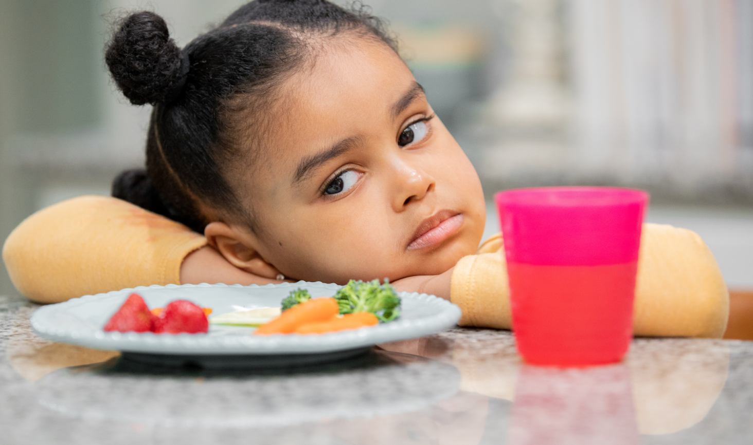 Young girl rests her chin on her arms at the table, looking away from a plate of vegetables and fruit.