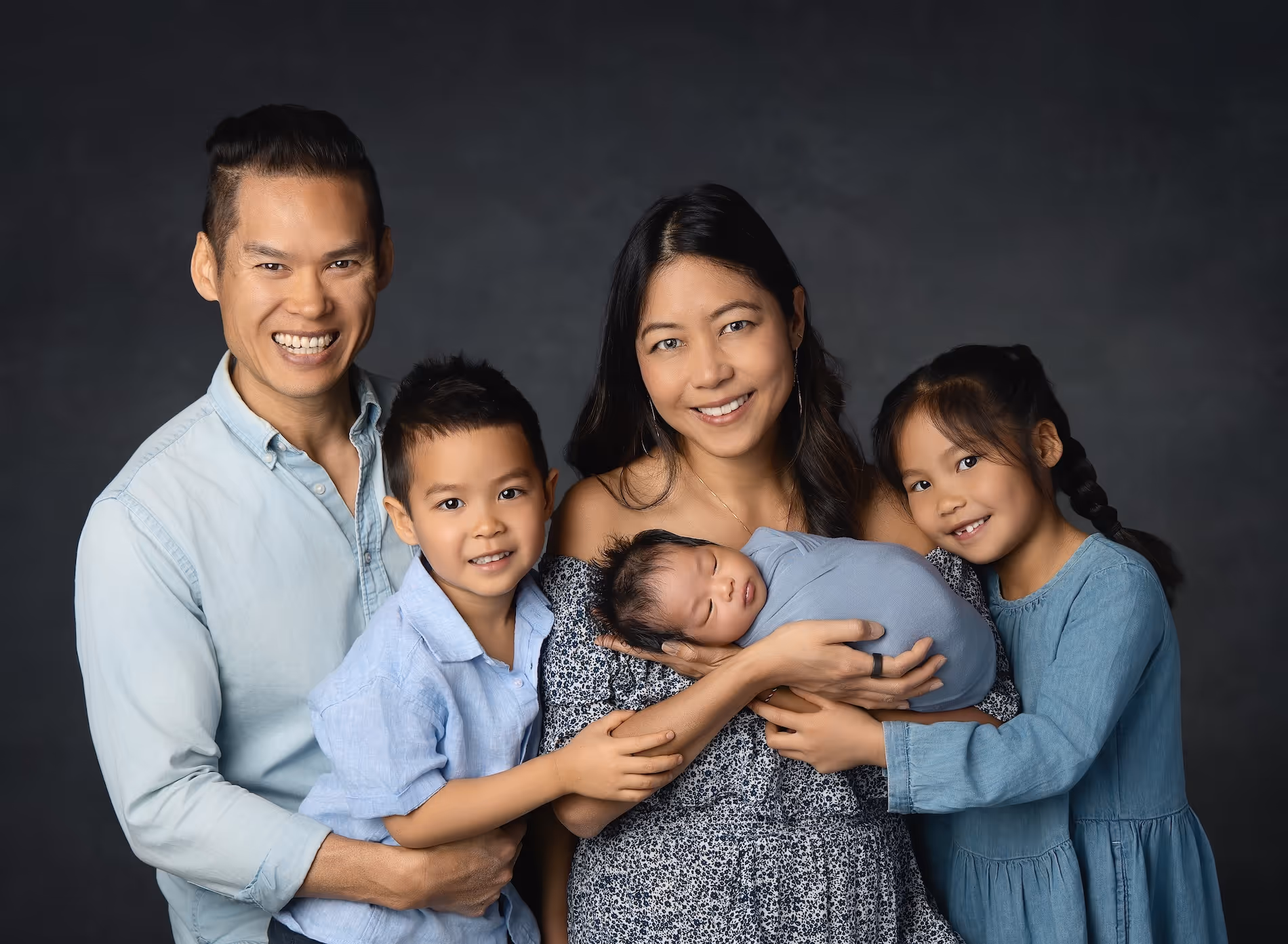 Cheryl Sew Hoy and her family of five pose with their swaddled newborn with a dark backdrop.