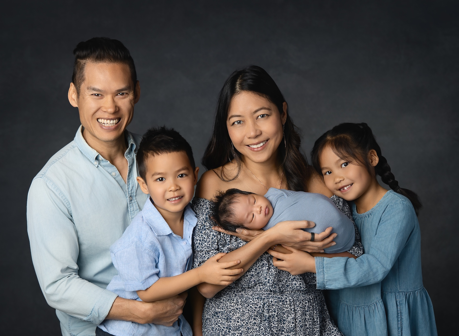 Cheryl Sew Hoy and her family of five pose with their swaddled newborn with a dark backdrop.