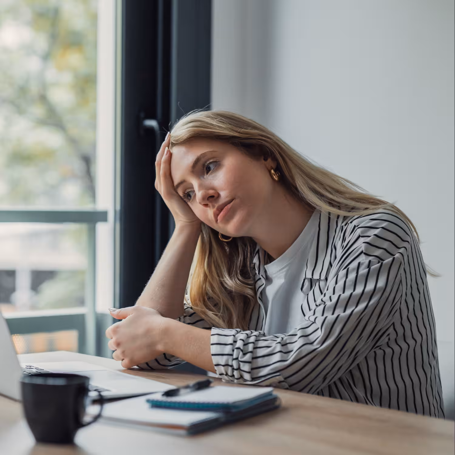 Tired woman resting her head on her hand while sitting at a desk with a laptop and coffee, looking fatigued and unfocused
