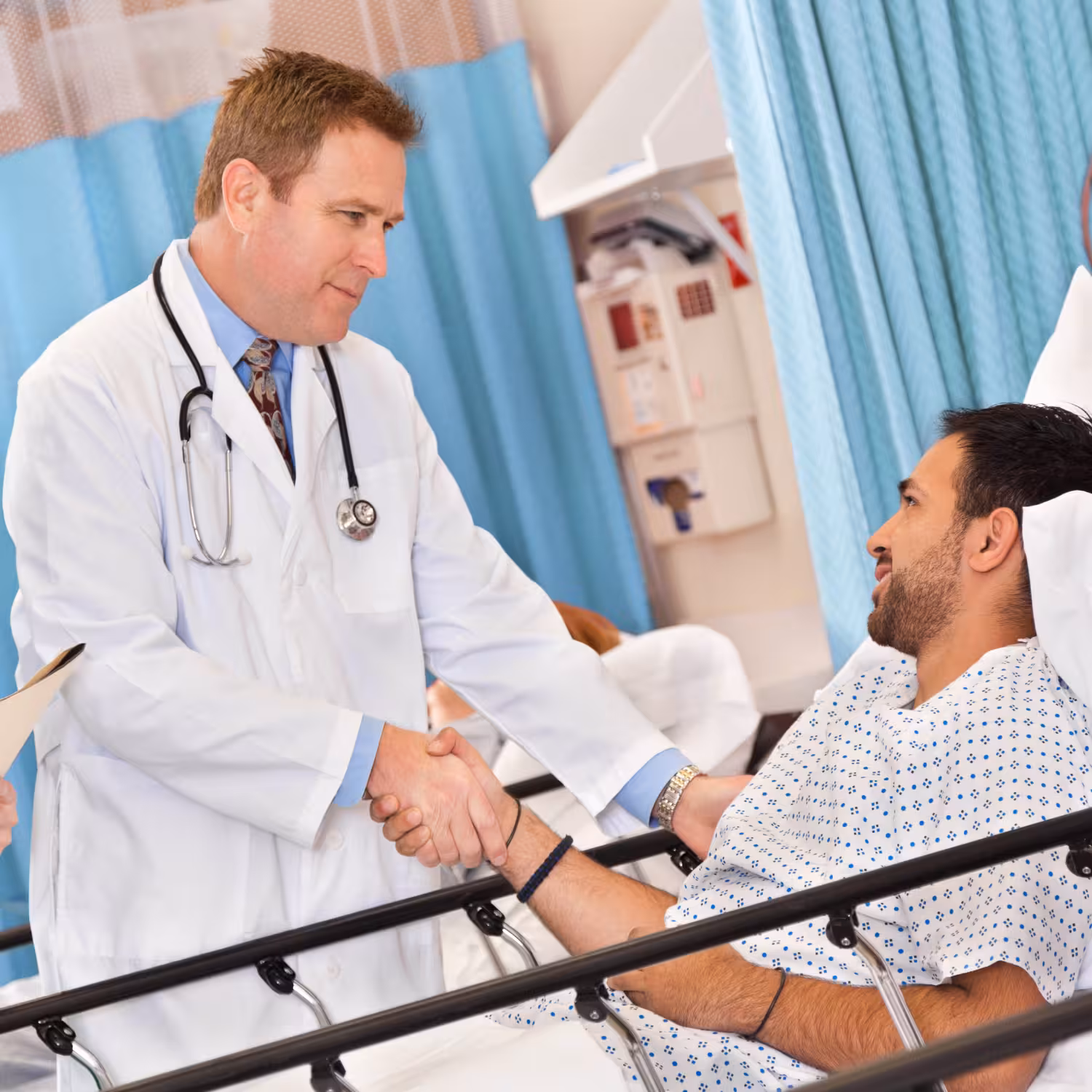 A doctor shaking hands with a hospital patient sitting in bed, both smiling, in a curtained hospital room.
