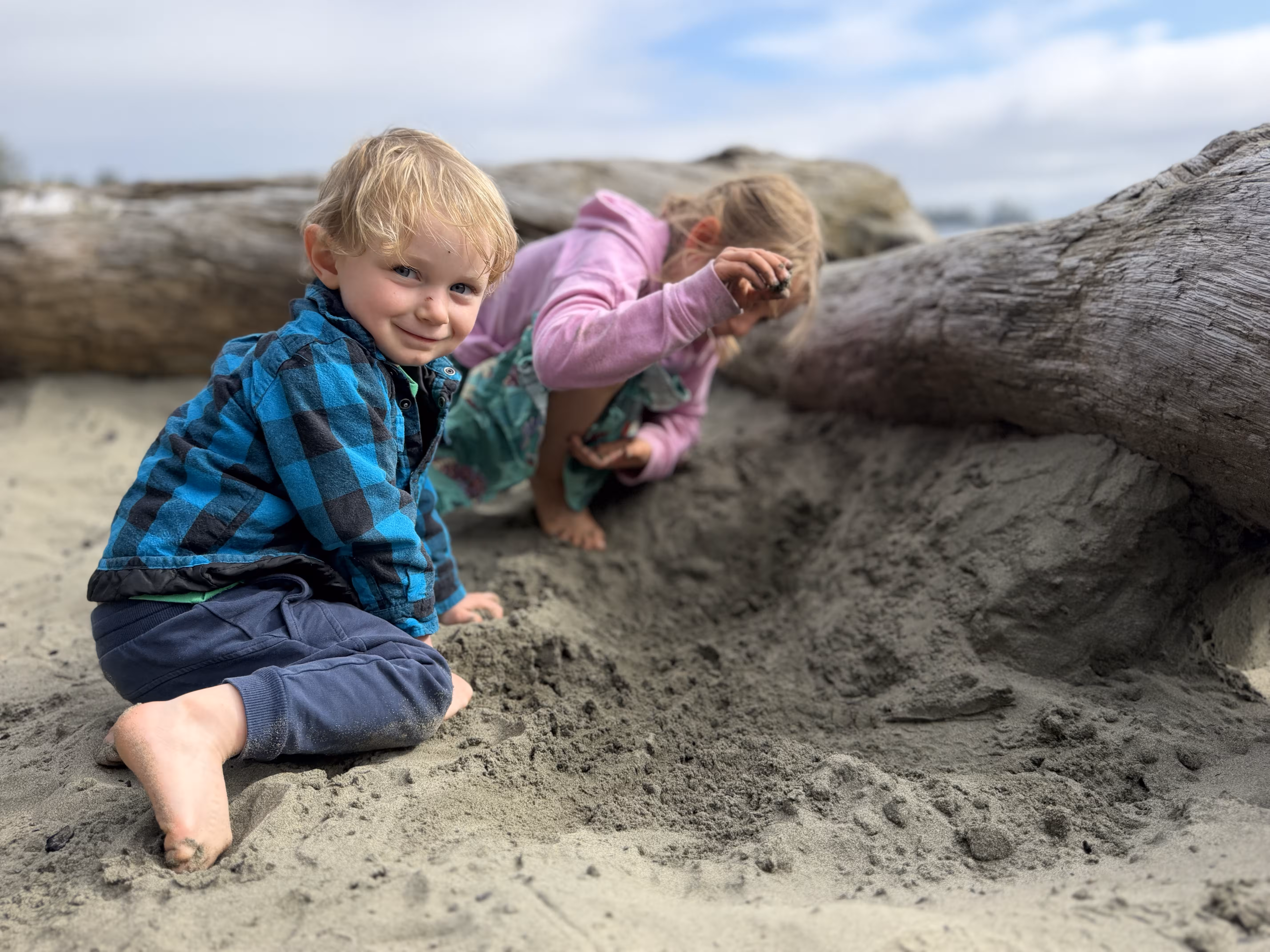 Blond toddler kneels in sand near driftwood, smiling, sibling digging in background.