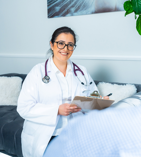 Female doctor with glasses and a stethoscope smiling and holding a clipboard while sitting in a cozy medical office.