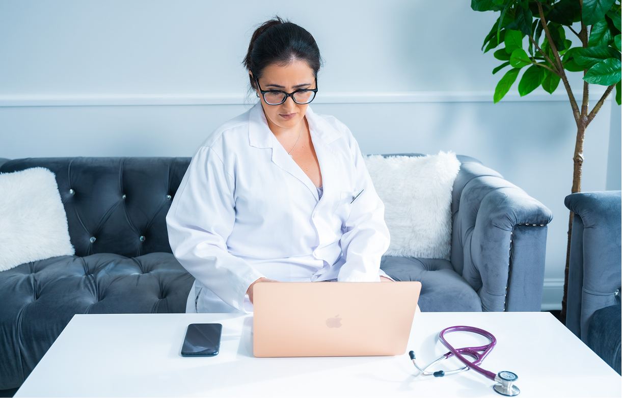 Female doctor in a white coat working on a laptop at a white table with a smartphone and stethoscope nearby, seated on a gray sofa.