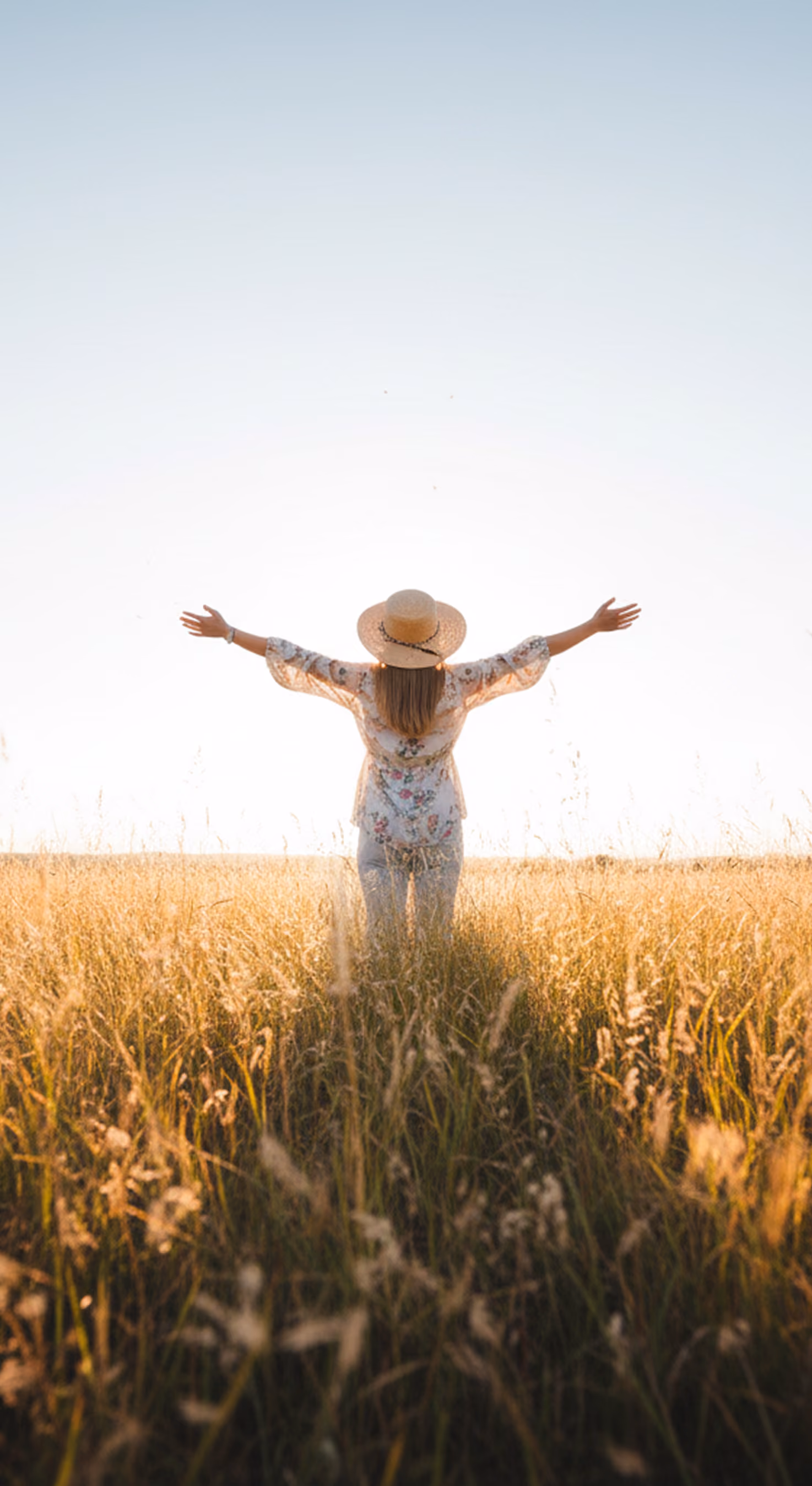Person in a floral shirt and hat with arms wide open standing in a sunlit field of tall grass.