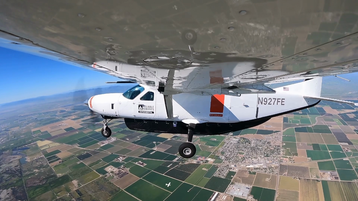A white single-engine aircraft flying above patchwork farmland.