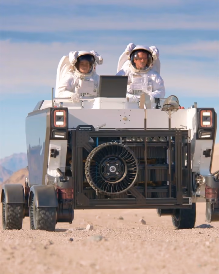Two suited astronauts riding an Astrolab FLEX rover across desert terrain
