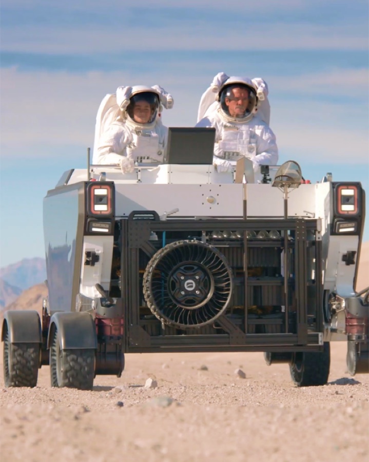 Two suited astronauts riding an Astrolab FLEX rover across desert terrain