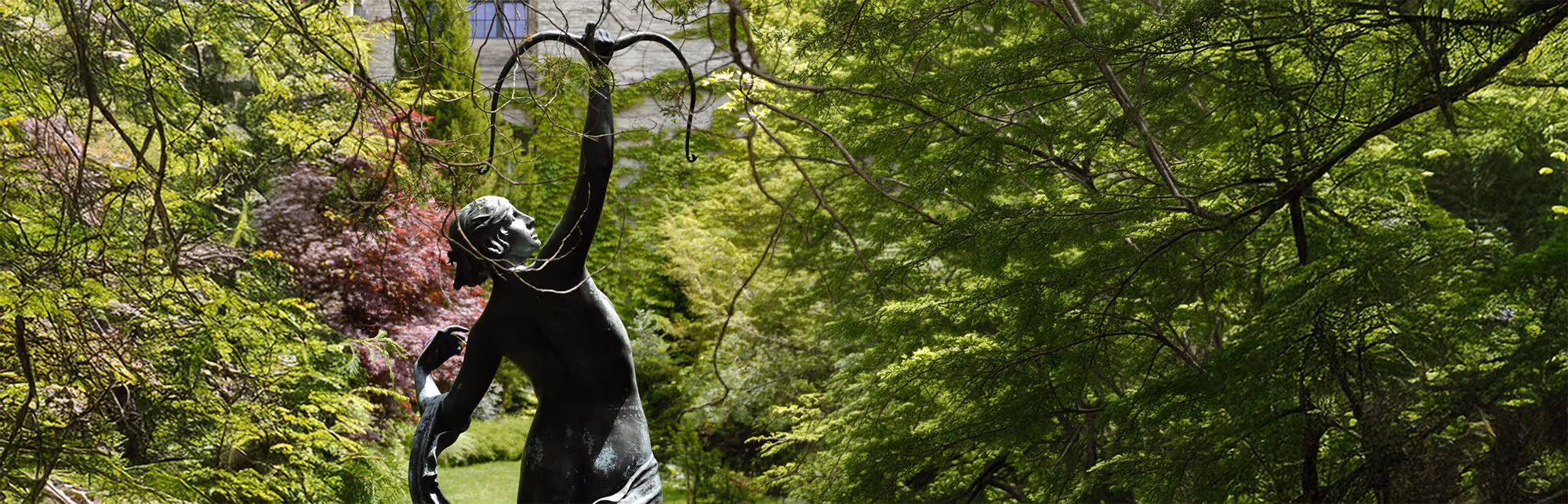 Bronze statue of a woman with arms raised, surrounded by lush green trees in a garden.