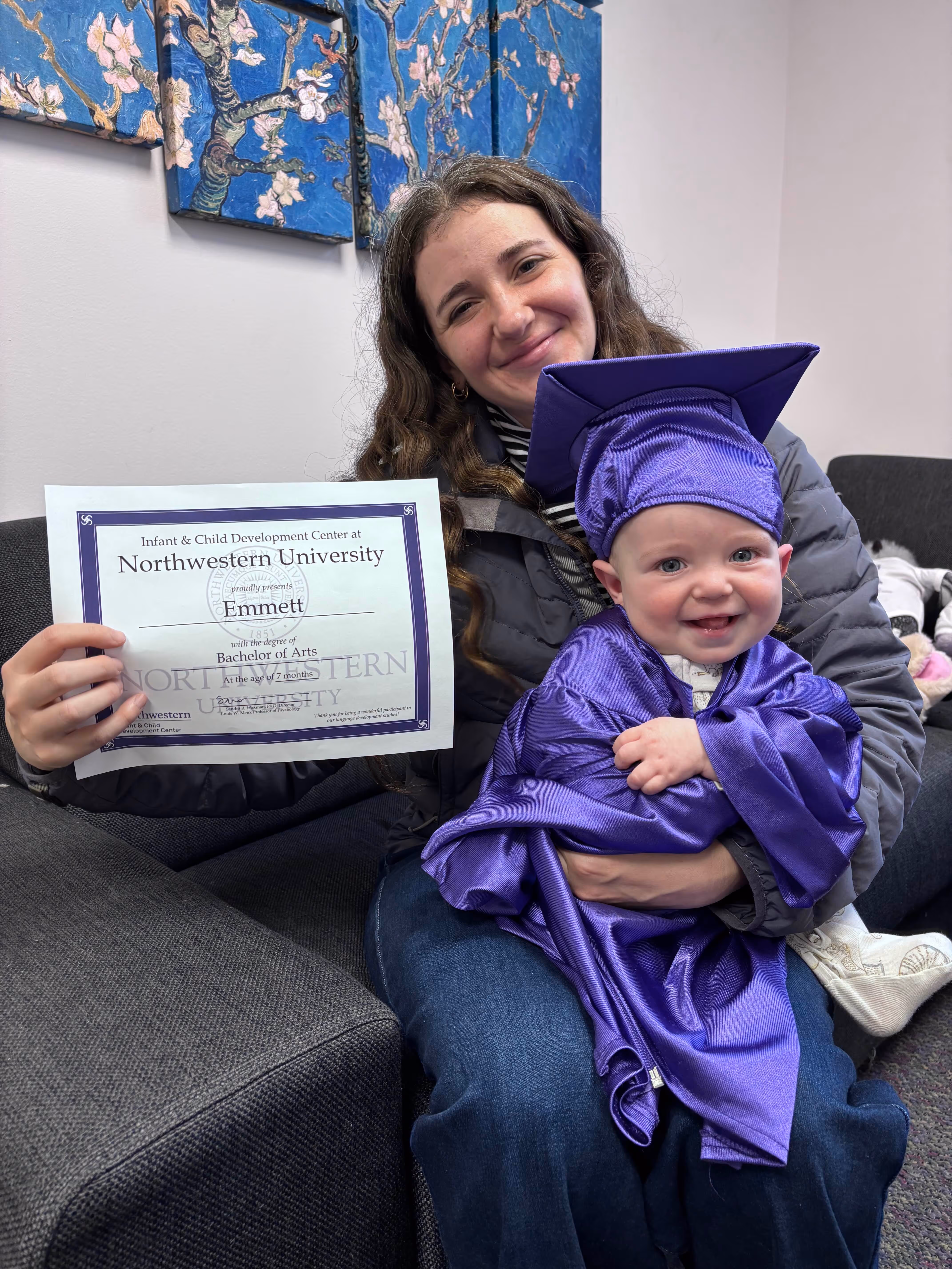 Woman holding a smiling baby dressed in a purple graduation cap and gown sitting on a gray couch.