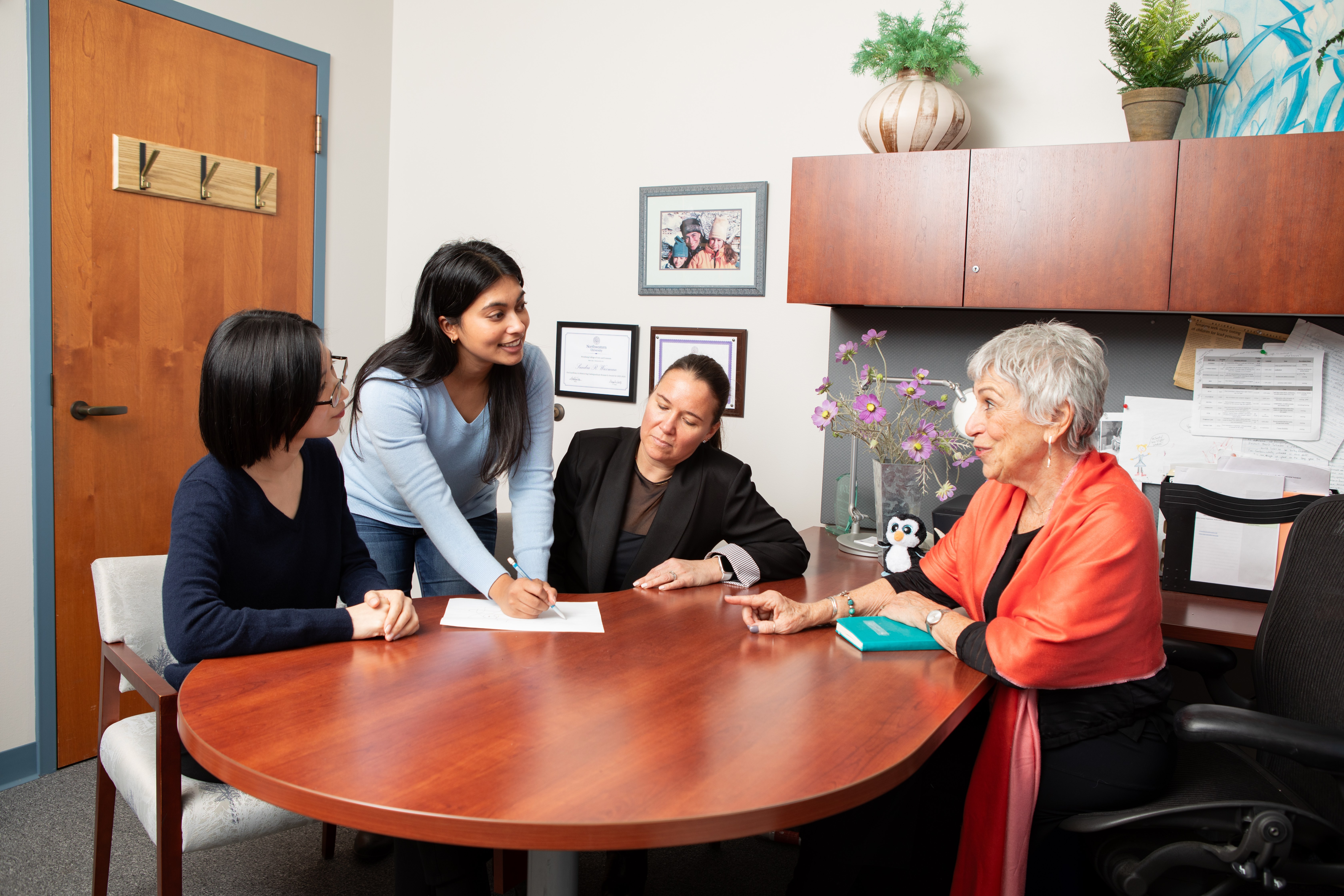 Four women gathered around a wooden office table, one standing and writing on paper.