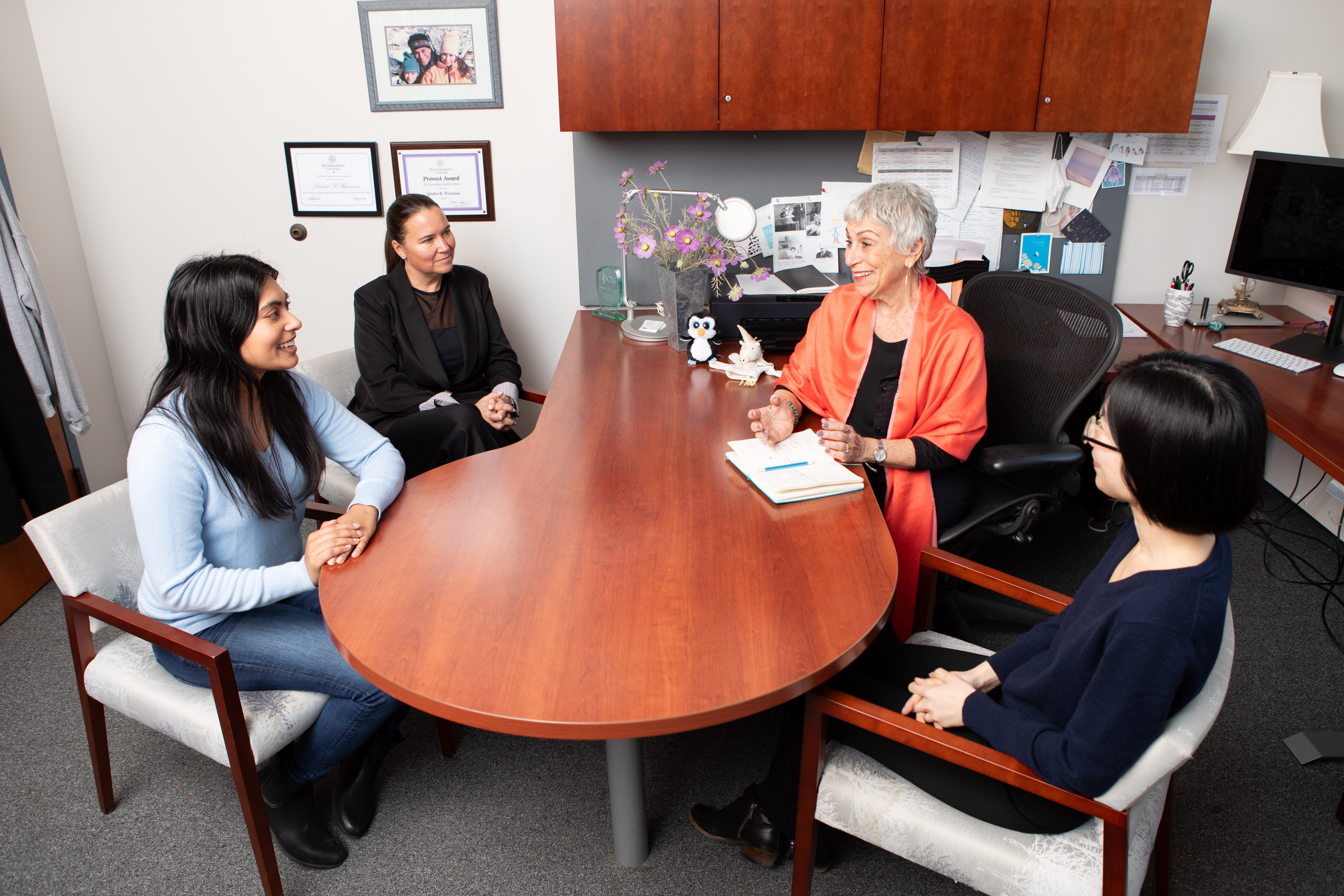 Four women engaged in a discussion around a wooden office desk in a professional setting.