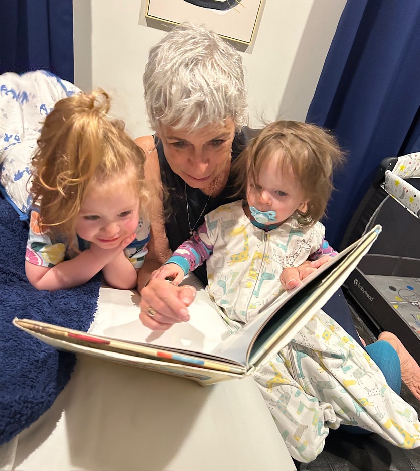 Older woman reading a book to two young children sitting closely together on a bed.