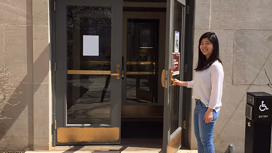 Woman with long dark hair wearing a white sweater and jeans opening a glass door of a building.