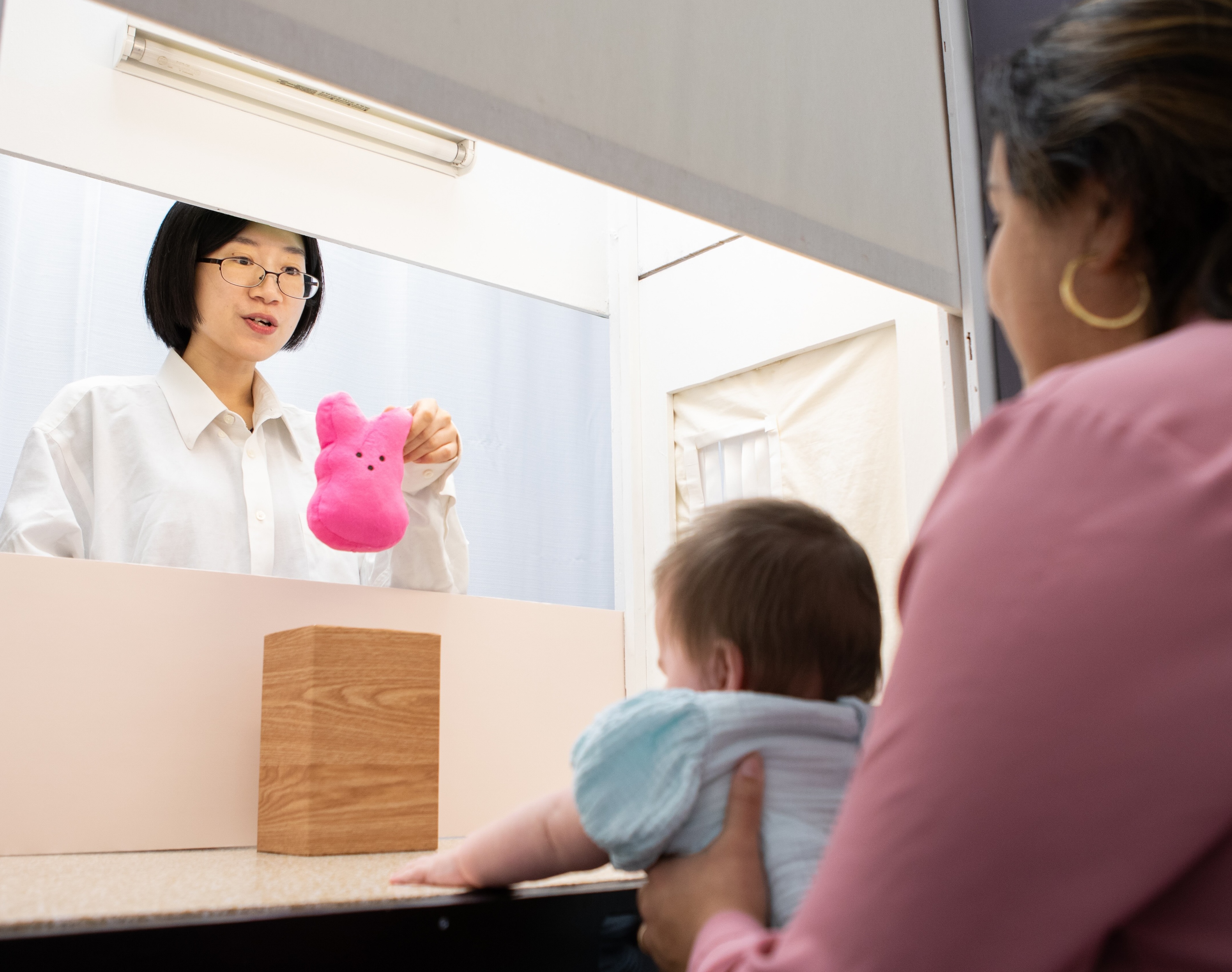 Woman in glasses showing a pink plush toy through a window to a baby held by another woman.