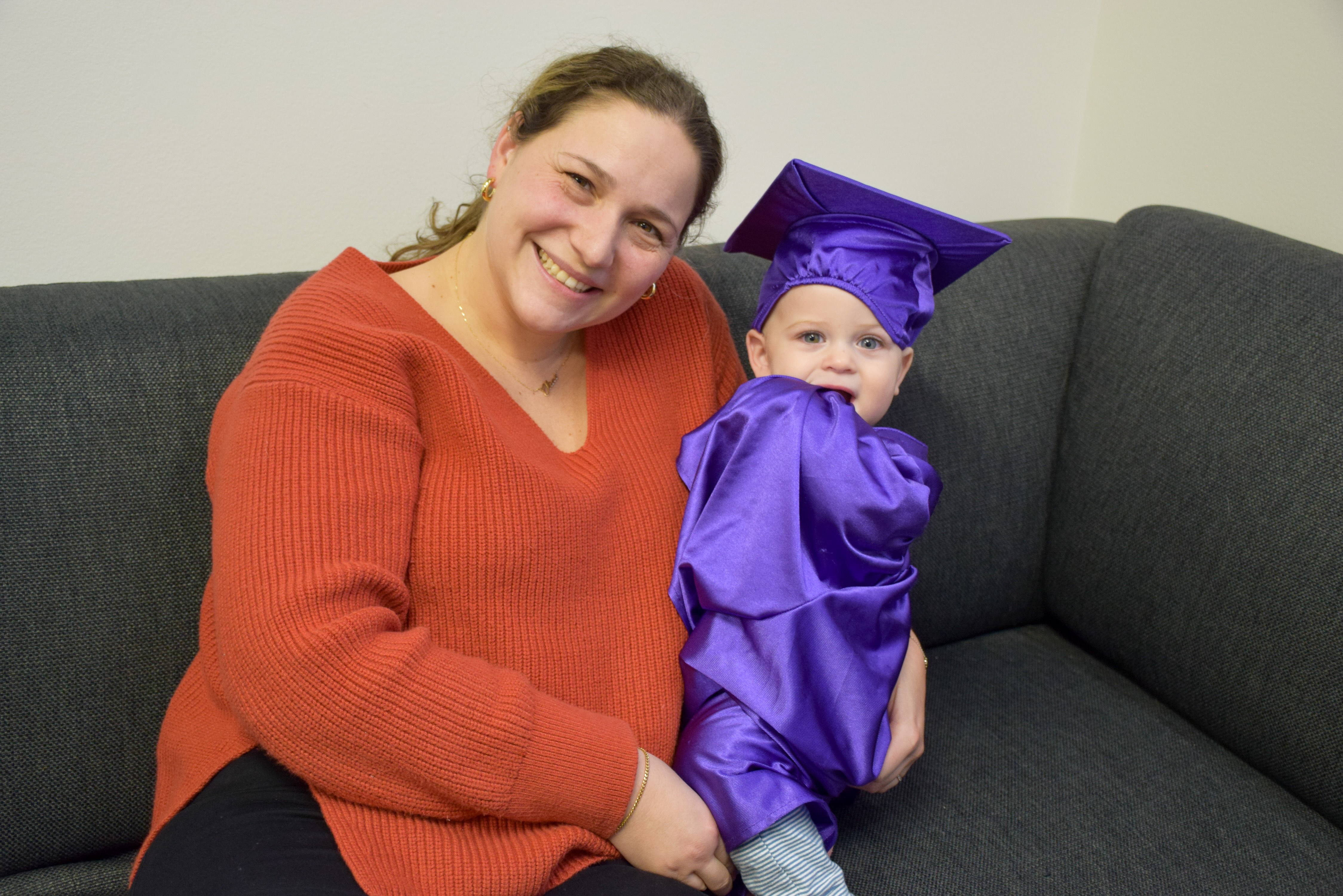 Smiling woman in orange sweater sitting on a gray couch holding a baby in a purple graduation gown.
