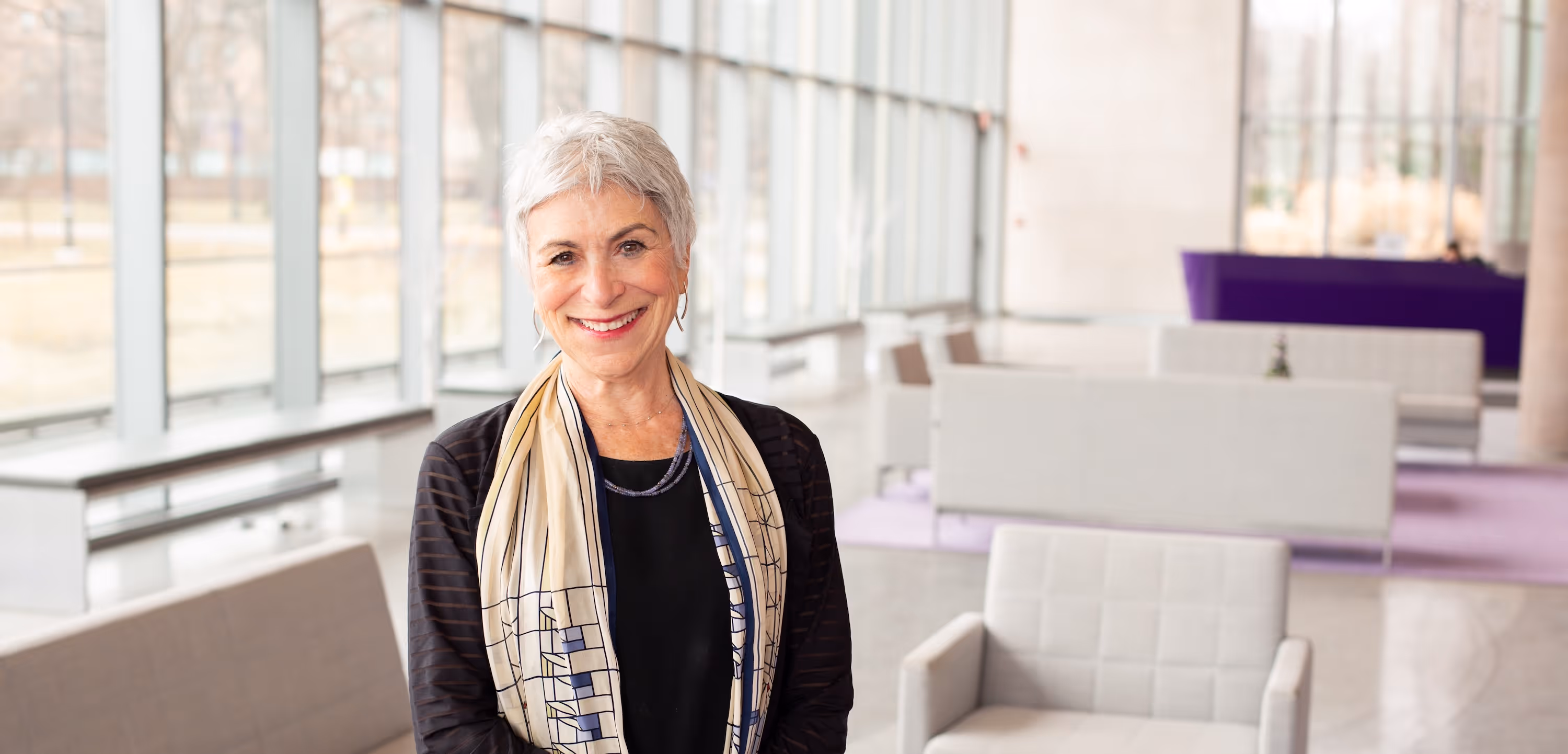 Smiling mature woman with short gray hair wearing a patterned scarf and black top in a modern lounge area.