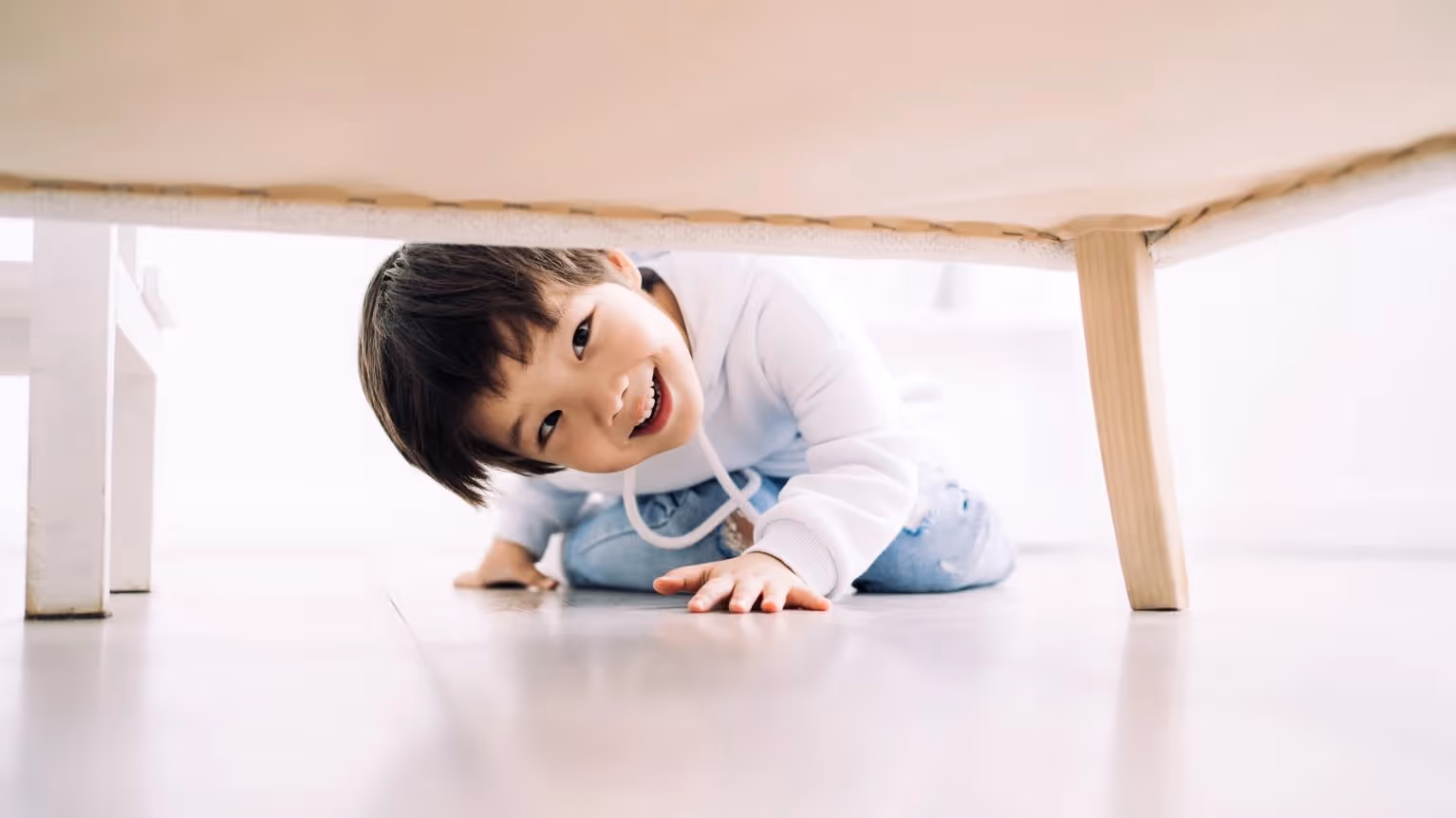 Smiling young boy crawling under a table on a bright floor.