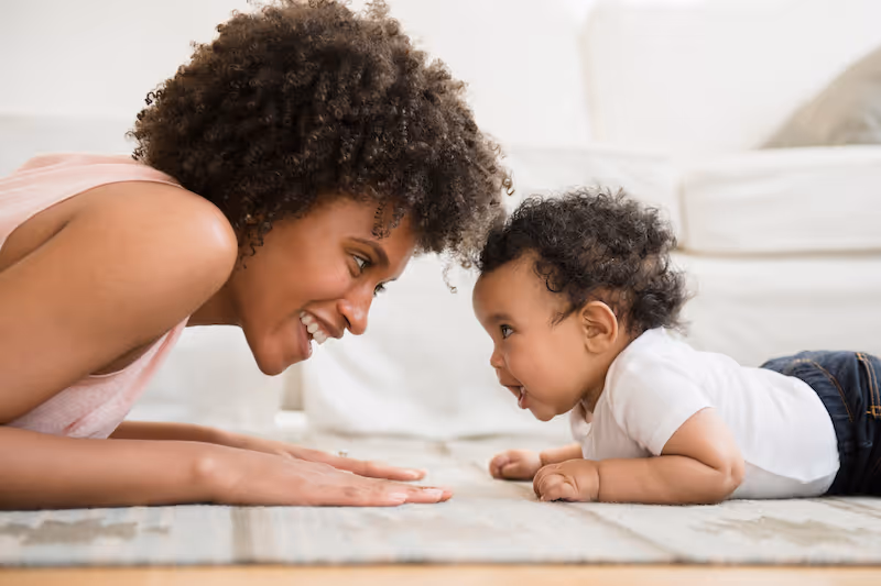 Woman and baby lying face to face on a rug, smiling and engaging closely indoors.