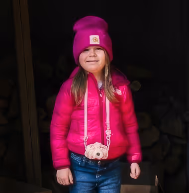 Young girl wearing a bright pink jacket and matching beanie with a small teddy bear purse standing against a dark background.