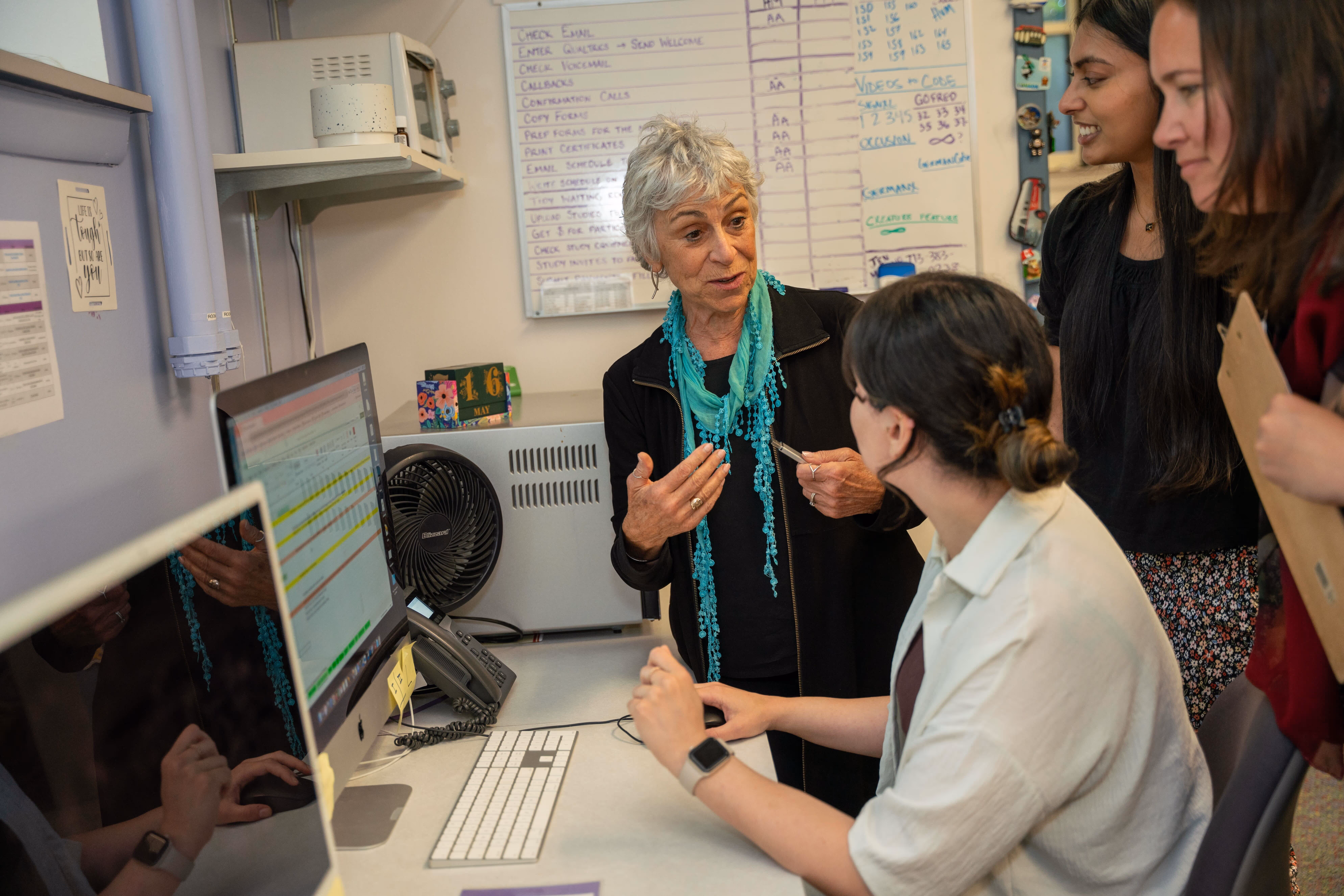 Sandy talking to seated young woman at a computer, two other women watch nearby.