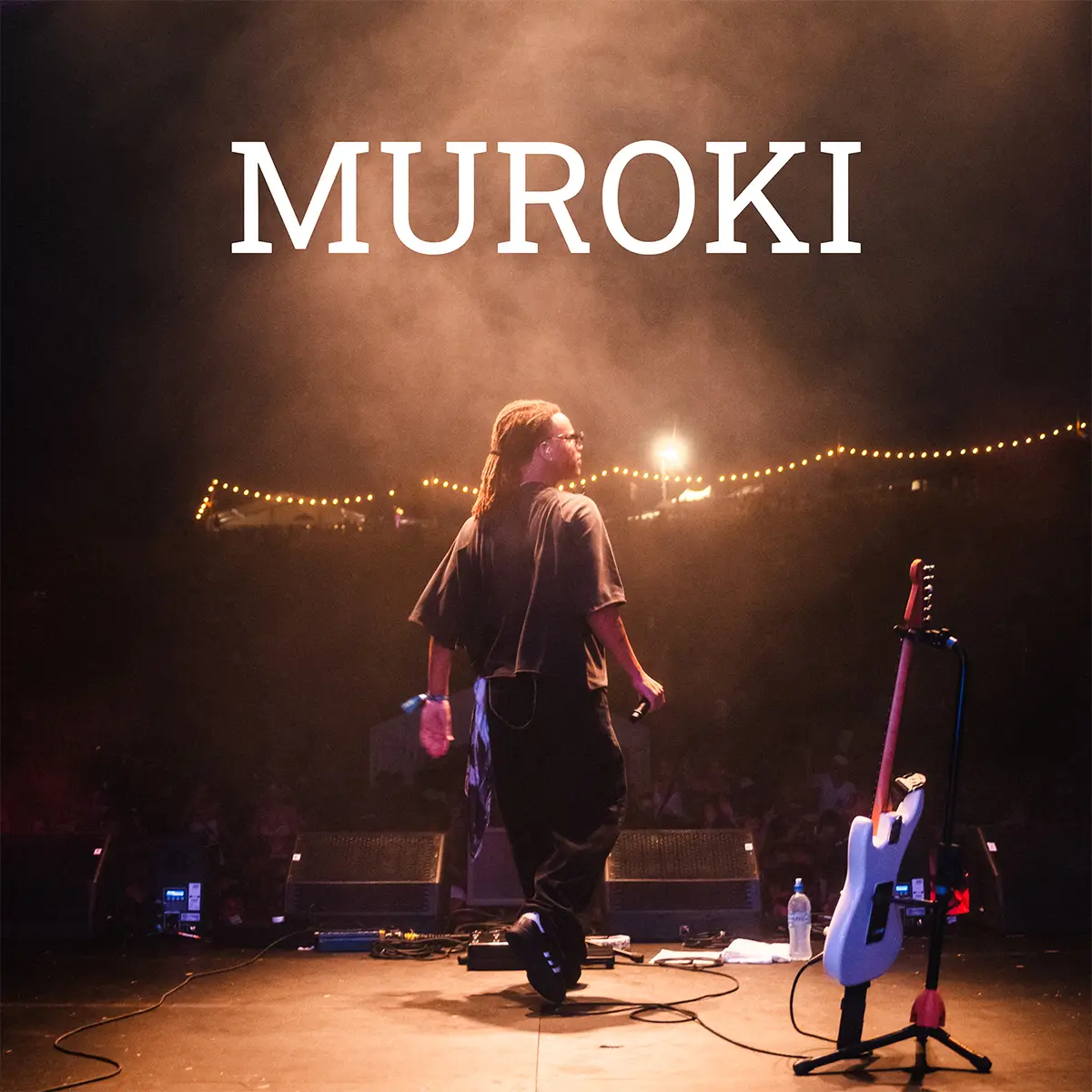 Muroki music performer with dreadlocks holding a microphone on stage with a guitar on a stand and string lights in the background.