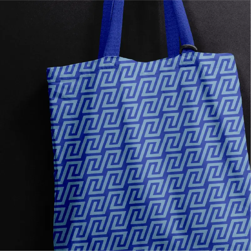 Blue tote bag with a darker blue geometric maze brand pattern and matching blue handles against a dark background.