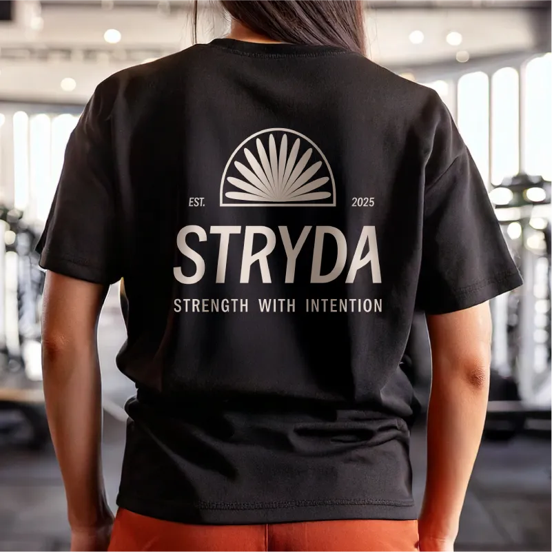 Woman wearing a black merch t-shirt with a design of the STRYDA logo and the text 'EST. 2025 STRENGTH WITH INTENTION' on the back in a STRYDA gym.