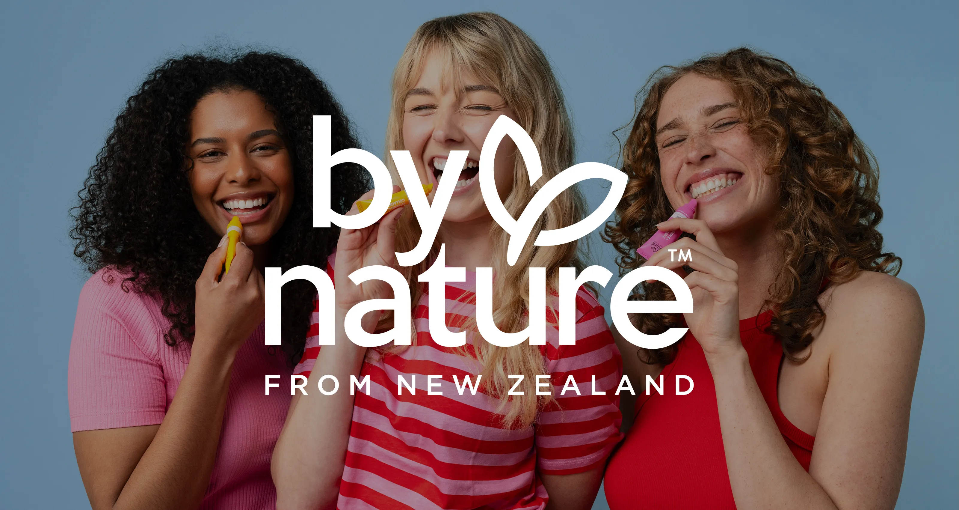 Three unique smiling women applying By Nature lip balms against a blue background with By Nature From New Zealand logo overlayed.