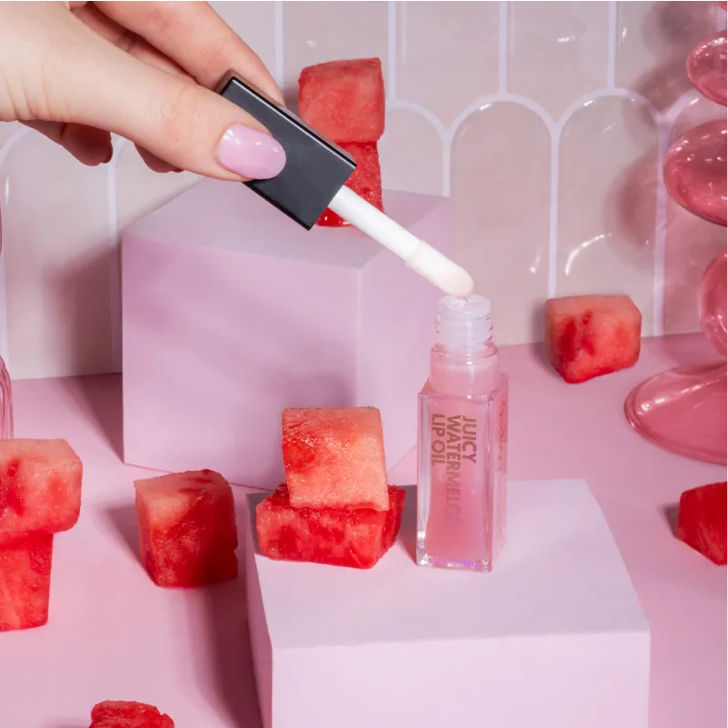 Hand with pink-painted nails holding a lip oil applicator over a clear pink By Nature Juicy Watermelon Lip Oil bottle surrounded by watermelon cubes on pastel pink blocks.