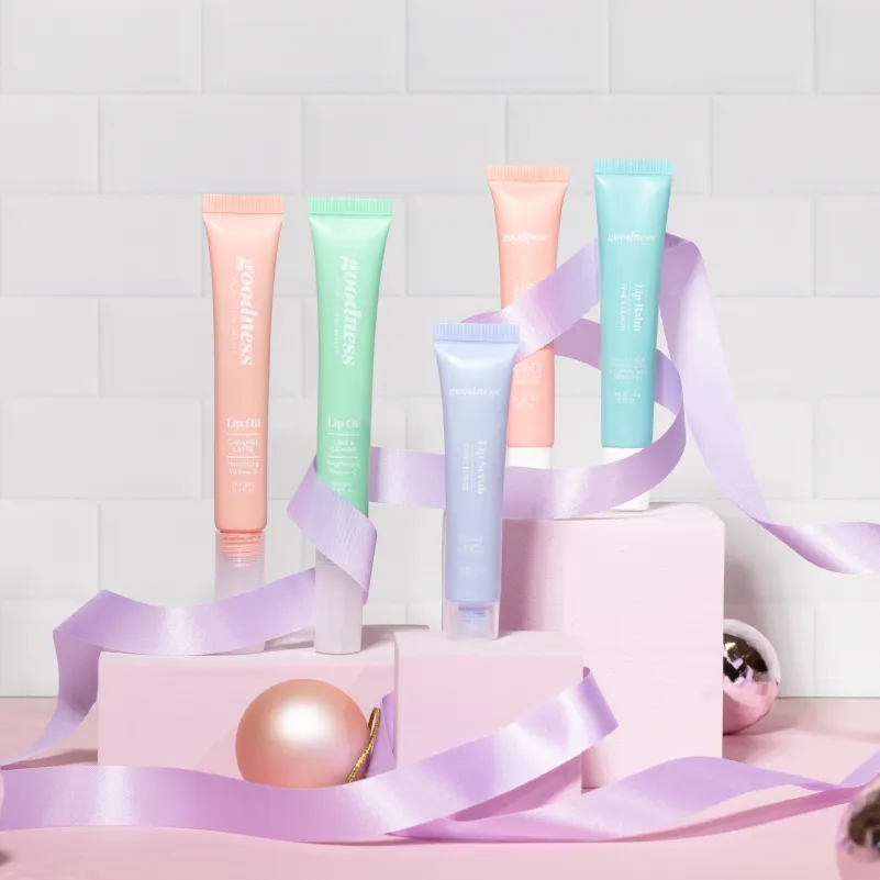 Five pastel-colored Goodness lip balm tubes arranged on pink blocks with a lavender ribbon intertwining and decorative ornaments.