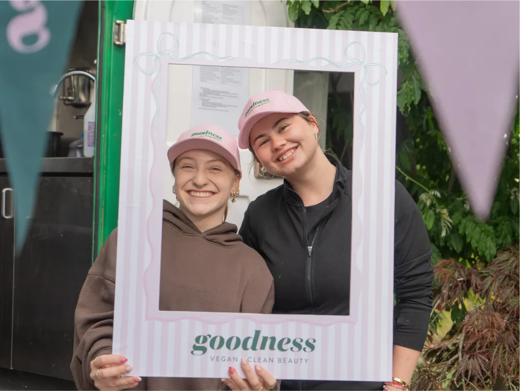 Samantha and employee from Gigi's Coffee and Matcha smiling holding a Goodness branded pink frame and wearing Goodness branded pink hats with flags in the foreground.