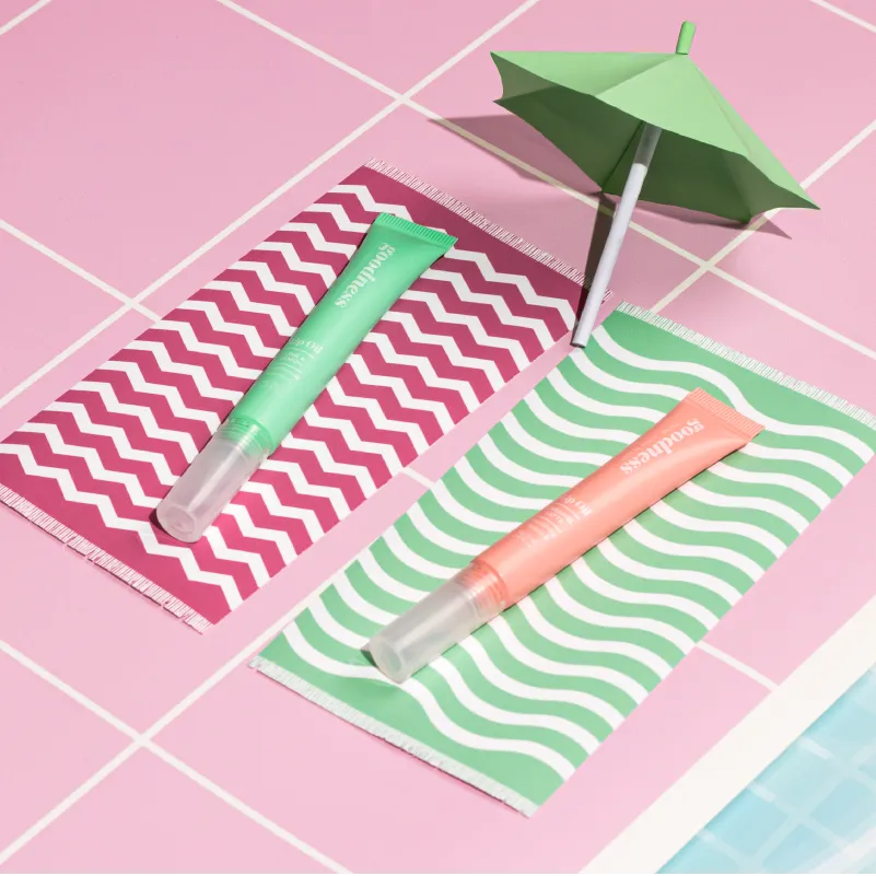 Two tubes of Goodness skincare lip oils placed on small colorful patterned towels beside a green paper umbrella on pink tiled surface next to a pool.