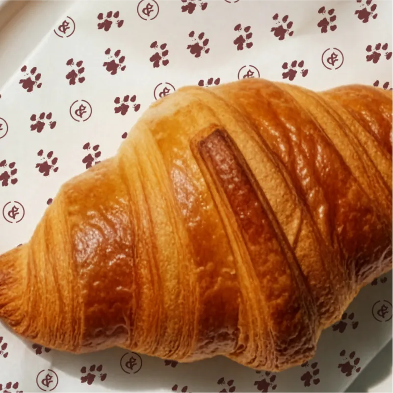 Close-up of a croissant on a branded Sip & Purr napkin featuring a custom paw print and cat-ear ampersand pattern.