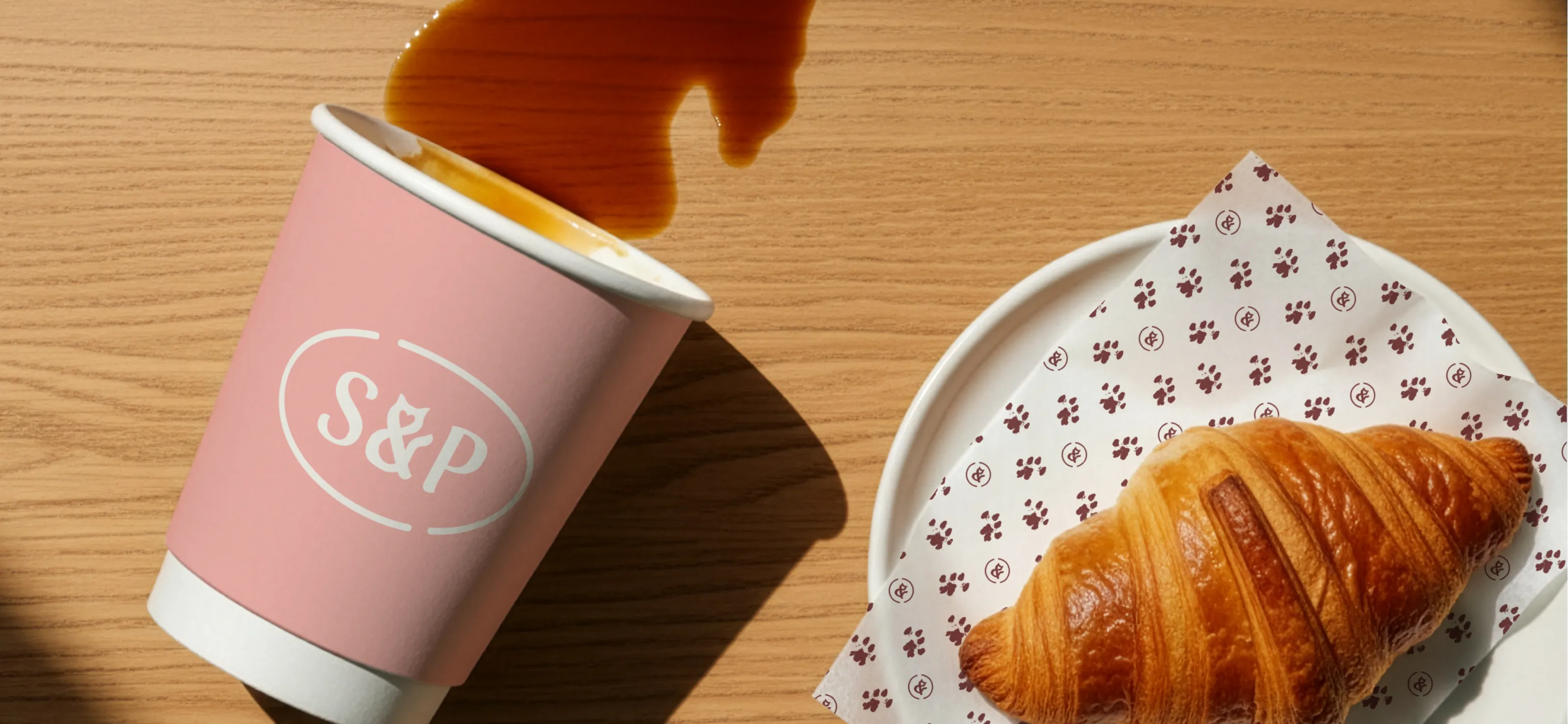 Graphic design for café hospitality packaging, featuring a croissant on custom brand pattern greaseproof paper and a branded takeaway coffee cup.
