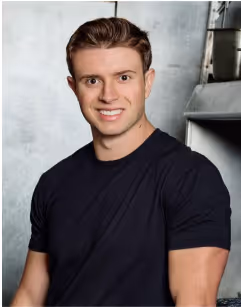 Young man with short brown hair wearing a black t-shirt, smiling against a light gray industrial background.