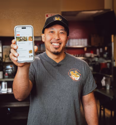 Smiling man in a restaurant uniform holding a smartphone displaying a menu app.