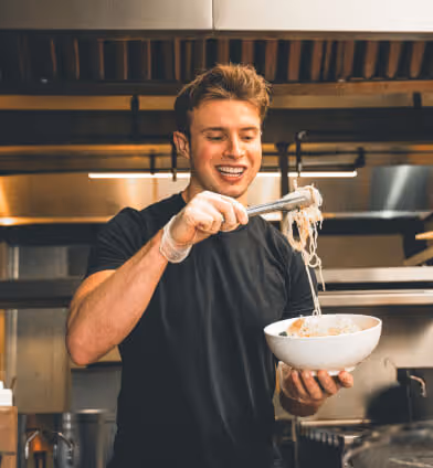Smiling young man using tongs to serve noodles into a white bowl in a kitchen.