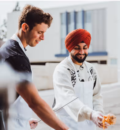 Two men wearing aprons preparing food together outdoors, one smiling and wearing a red turban.