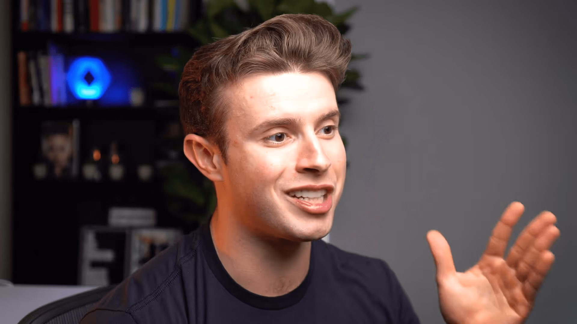 Young man with short brown hair speaking and gesturing with his right hand in an indoor setting.