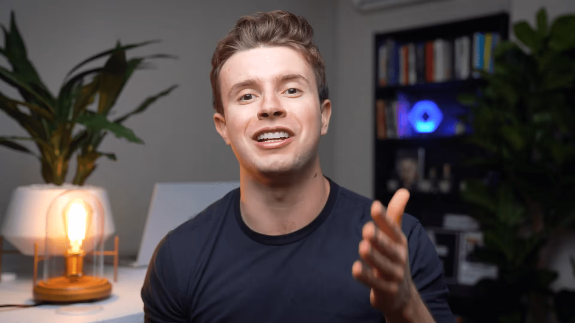 Young man with short brown hair wearing a black t-shirt, gesturing with his hand while speaking indoors.