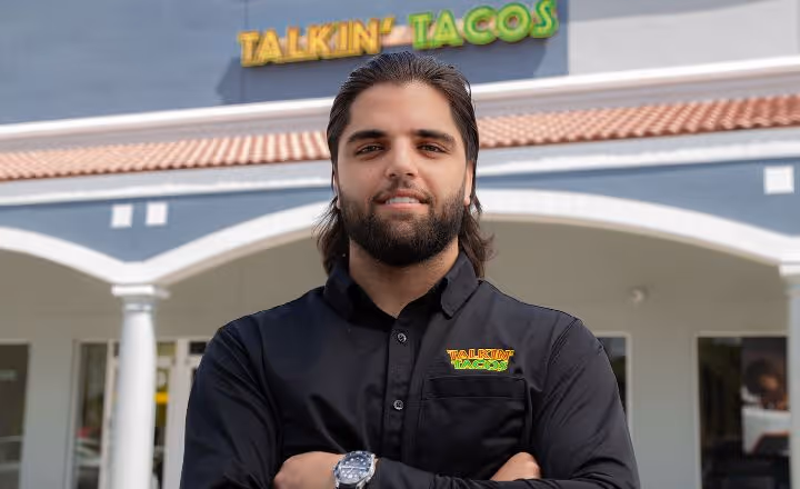 Man with dark hair and beard wearing a black Talkin’ Tacos shirt standing with arms crossed in front of the Talkin’ Tacos restaurant.